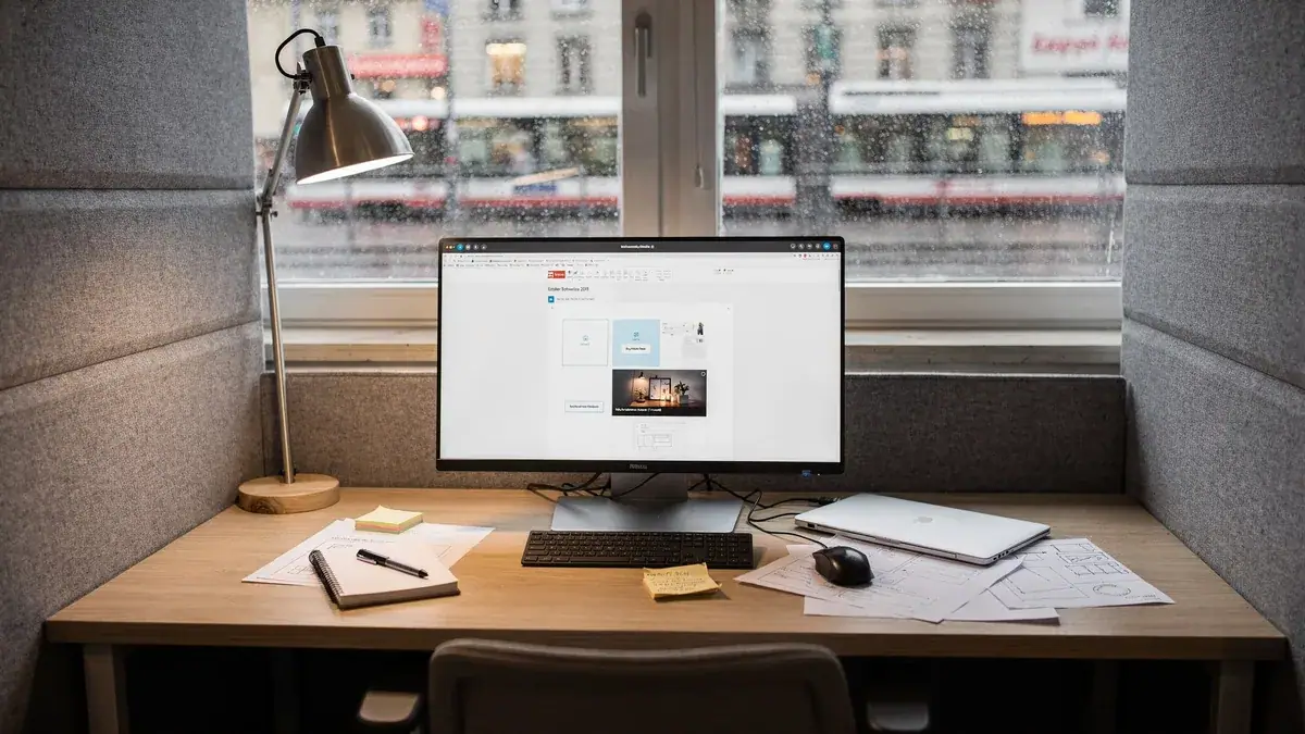 Monitor mit Website-Baukasten auf Holztisch am Fenster, Tischlampe, Laptop und handschriftliche Notizen.
